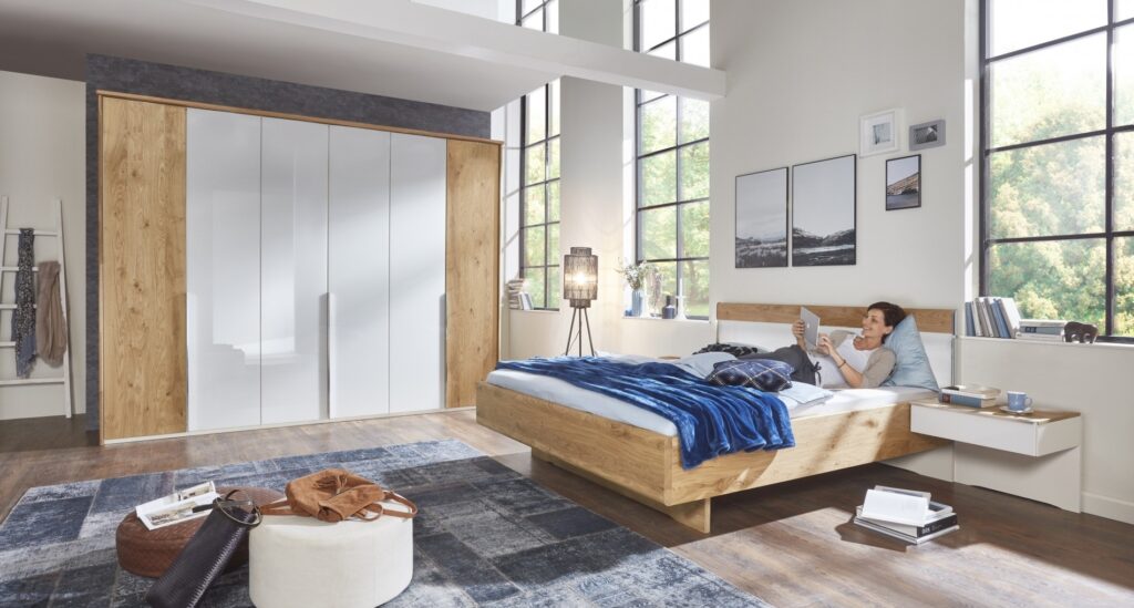 Modernes Schlafzimmer mit Holzbett und einem Holzschrank, eine Frau liegt in dem Bett und schaut etwas auf einem Tablet.