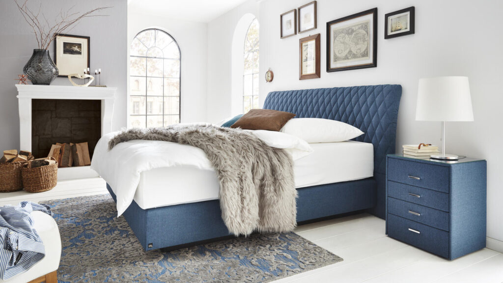 Blaues Polsterbett mit flauschiger Decke in einem Schlafzimmer mit Kamin und Wandkunst.