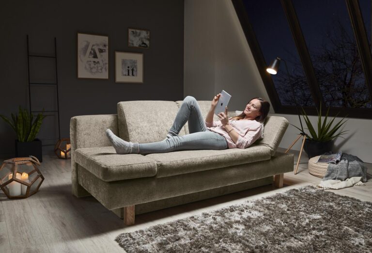 Frau entspannt sich auf einem beigen Sofa und benutzt nachts ein Tablet.