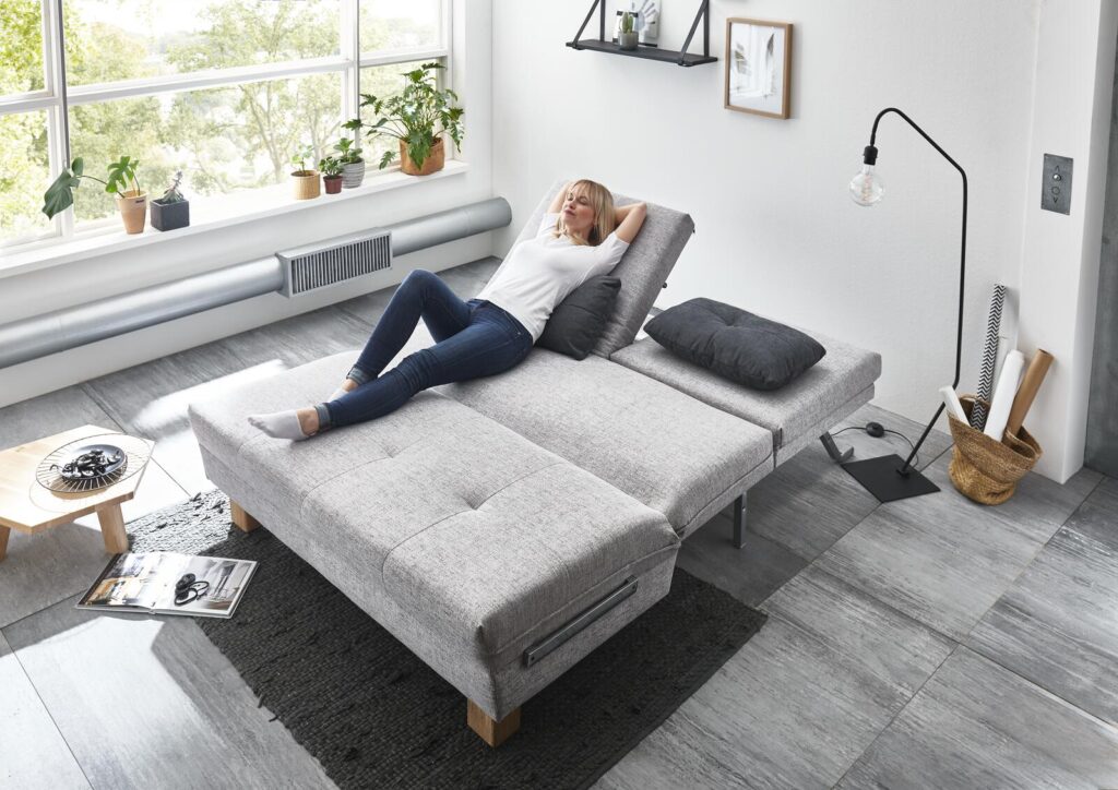 Frau entspannt sich auf einem grauen Stoffschlafsofa in einem modernen Wohnzimmer.