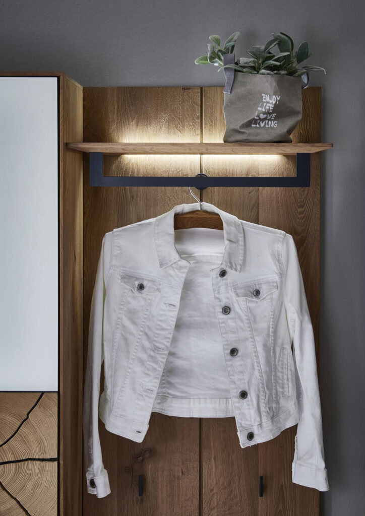Eine weiße Jeansjacke hängt an einem hölzernen Wandregal mit LED-Beleuchtung. Auf einem Blumentopf im Regal steht „Enjoy Life Love Living“.