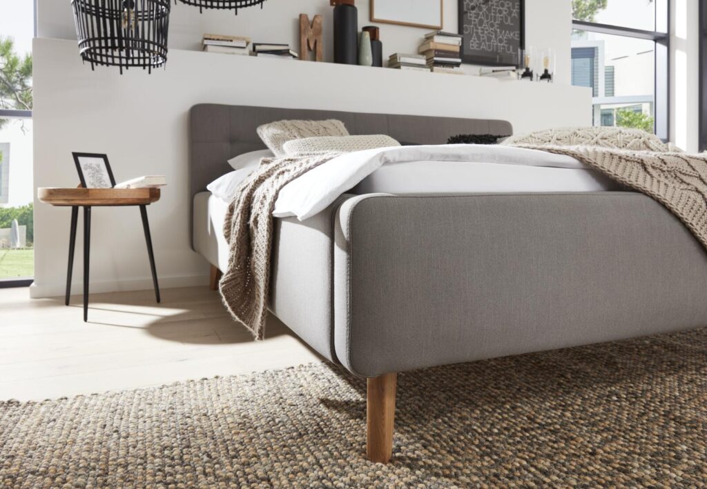 Grau gepolstertes Bett mit Holzbeinen, kuscheliger Strickdecke und Beistelltisch.