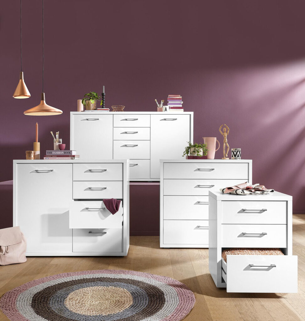 Weißes, modernes Schlafzimmermöbelset mit Kommode, Frisierkommode und Nachttisch vor einer malvenfarbenen Wand.