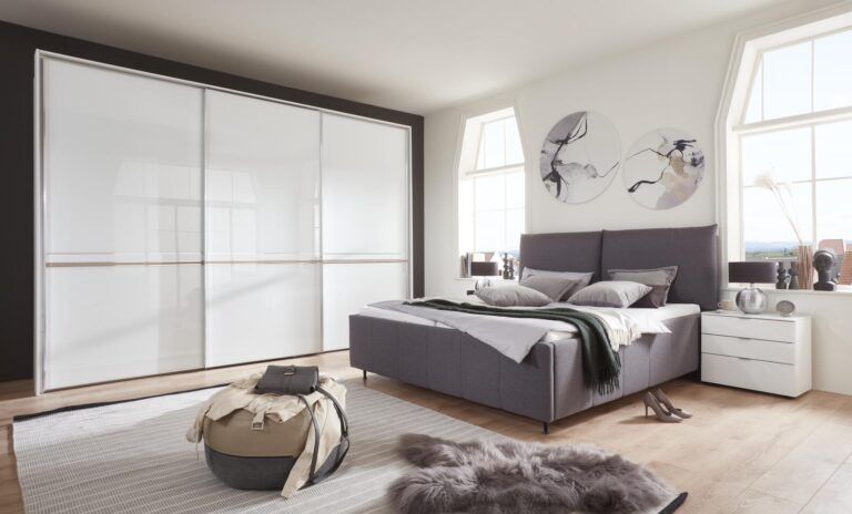 Modernes Schlafzimmer mit grauem Polsterbett, weißem Schiebetürenschrank und minimalistischem Nachttisch.