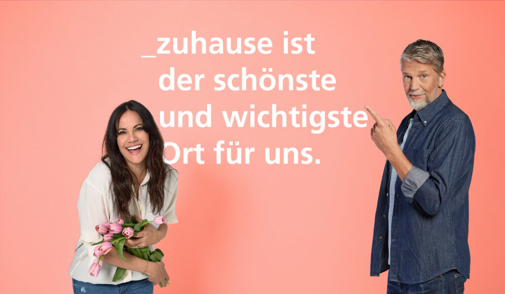 Eine Frau auf der linken Seite und ein Mann auf der rechten Seite, die Frau hält rosten Tulpen in der Hand und lacht in die Kamera, der Mann zeigt, während er in die Kamera schaut, auf den Text, der sagt "Zuhause ist der schönste und wichtigste Ort für uns"