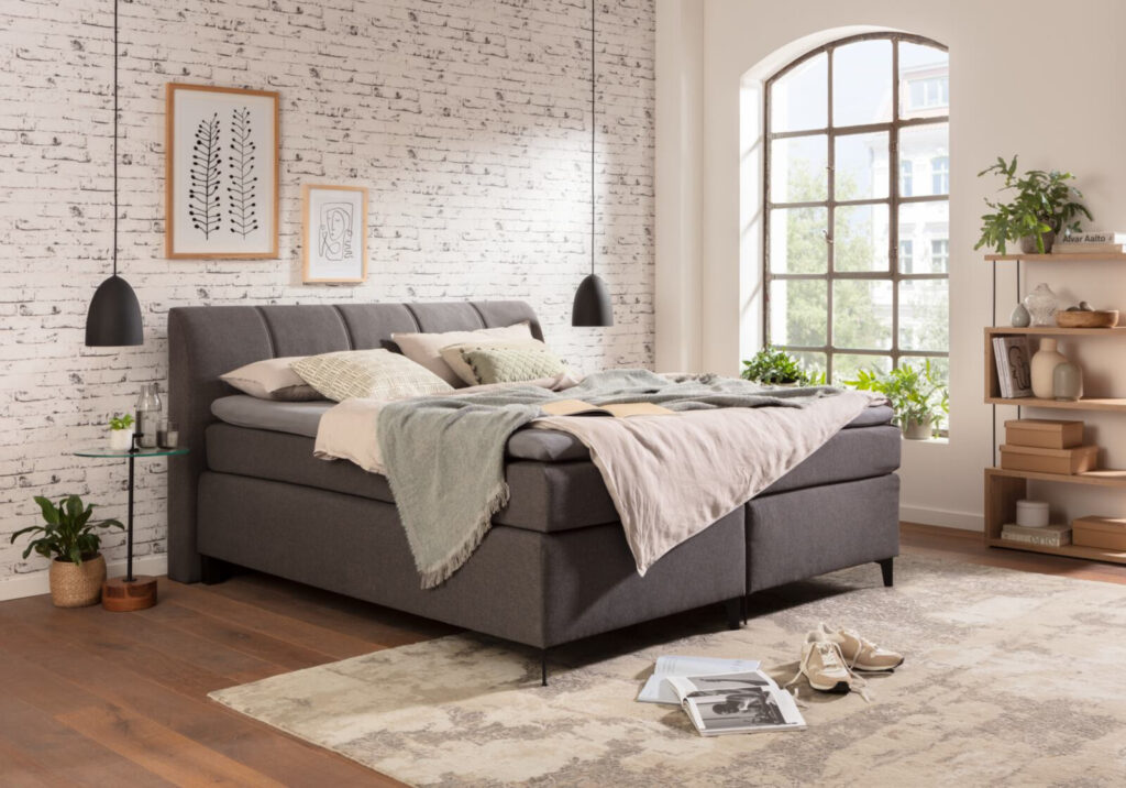 Graues Boxspringbett vor Backsteinwand mit Dekoration und großem Fenster.