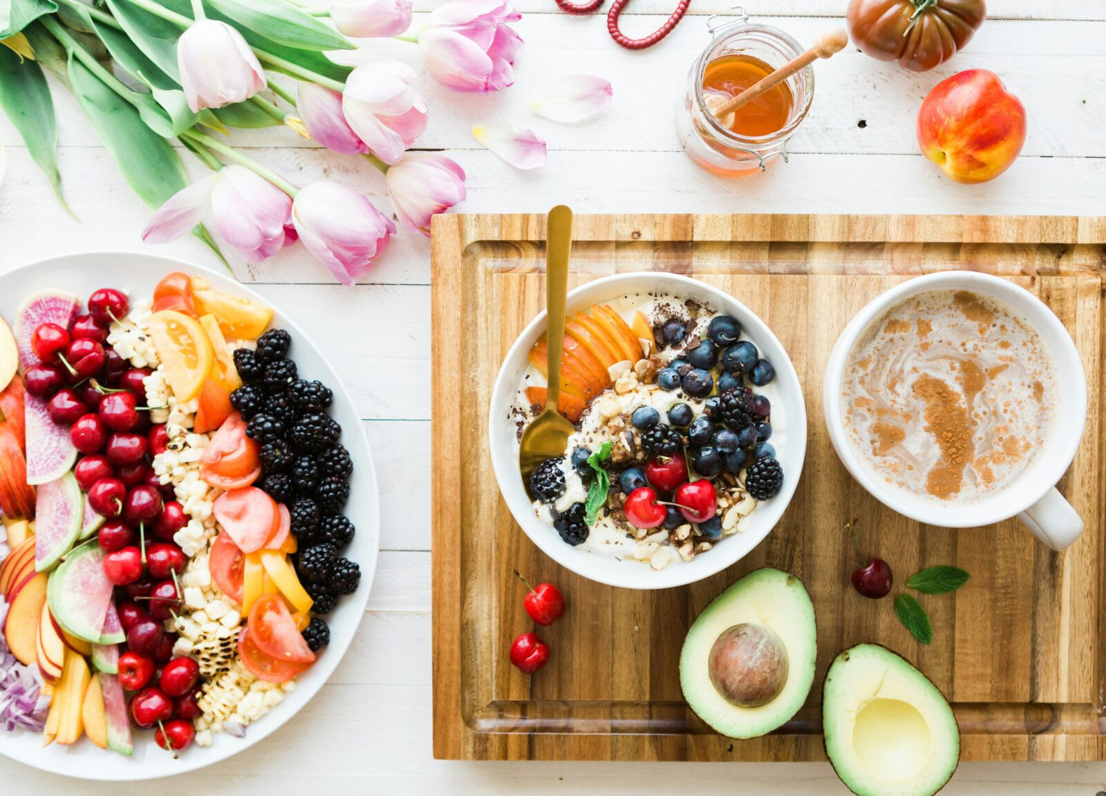 Gesunde Ernährung: Schüssel mit Obst, Beeren, Avocado, Kaffee und Blumen auf einem Holztisch.